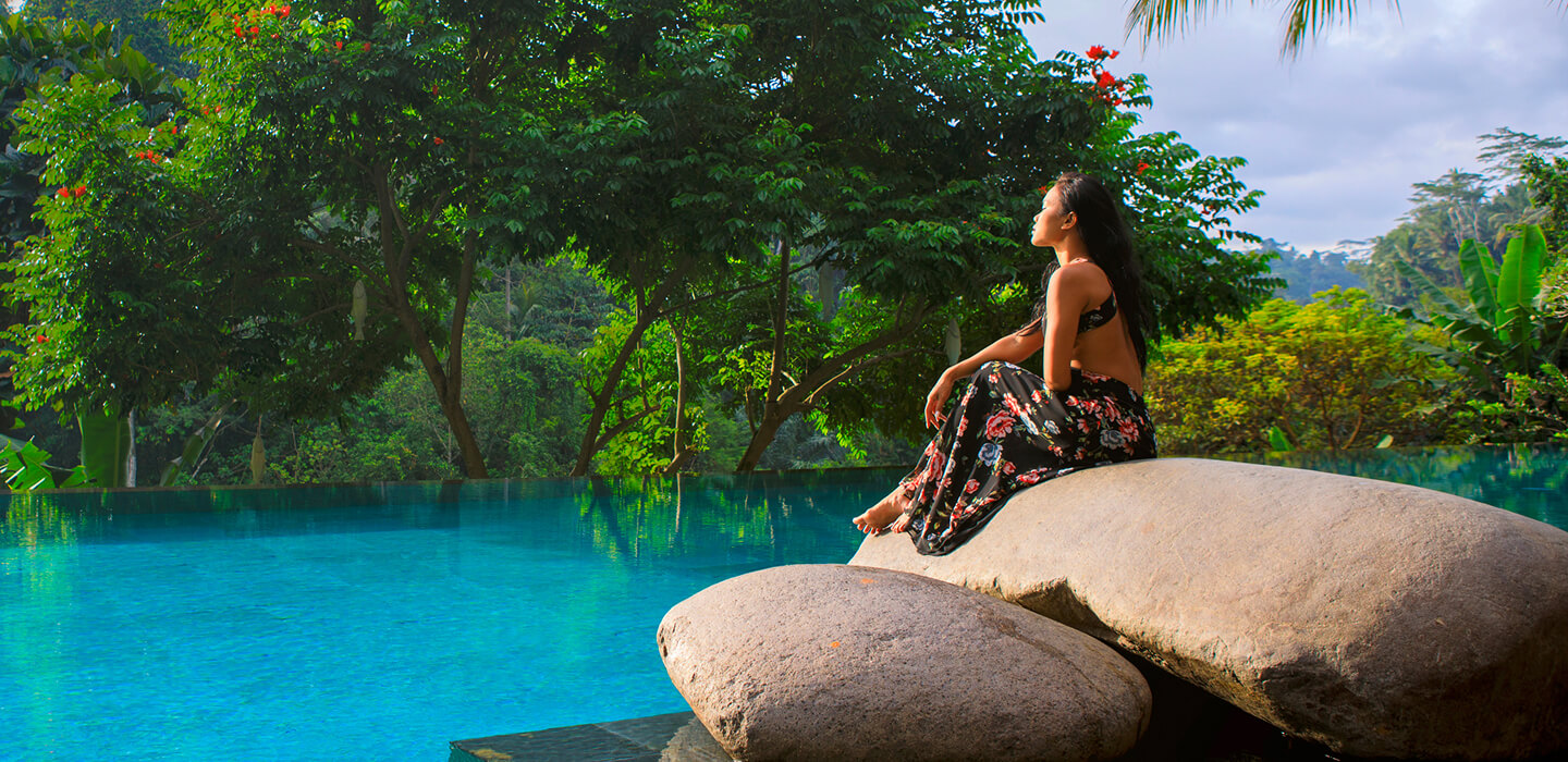 A woman sits on large rocks beside an infinity pool surrounded by lush tropical greenery.