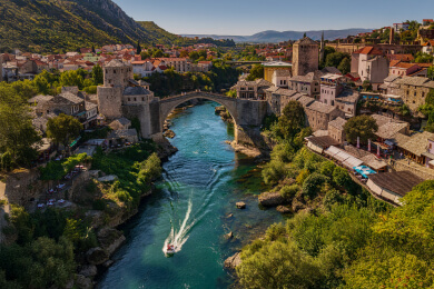 Scenic view of the historic Stari Most bridge in Mostar, Bosnia and Herzegovina, spanning the Neretva River, surrounded by stone buildings and lush green hills.