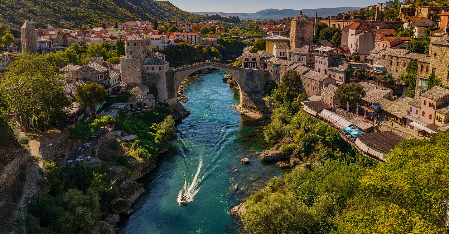 Scenic view of the historic Stari Most bridge in Mostar, Bosnia and Herzegovina, spanning the Neretva River, surrounded by stone buildings and lush green hills.